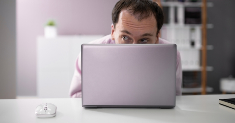 employee peeking out from behind his laptop, looking suspicious